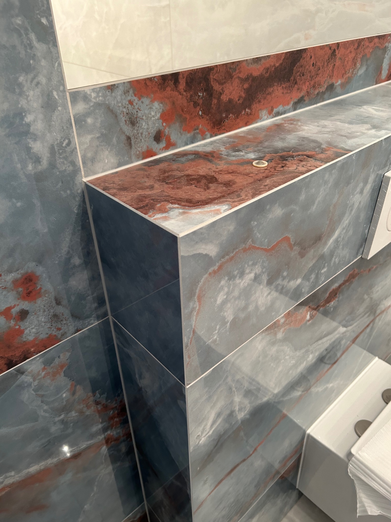 Półka w łazience wyłożona płytkami imitującymi marmur w odcieniach szarości i rdzawego brązu, z widocznym odpływem liniowym i fragmentem podajnika na papier toaletowy.