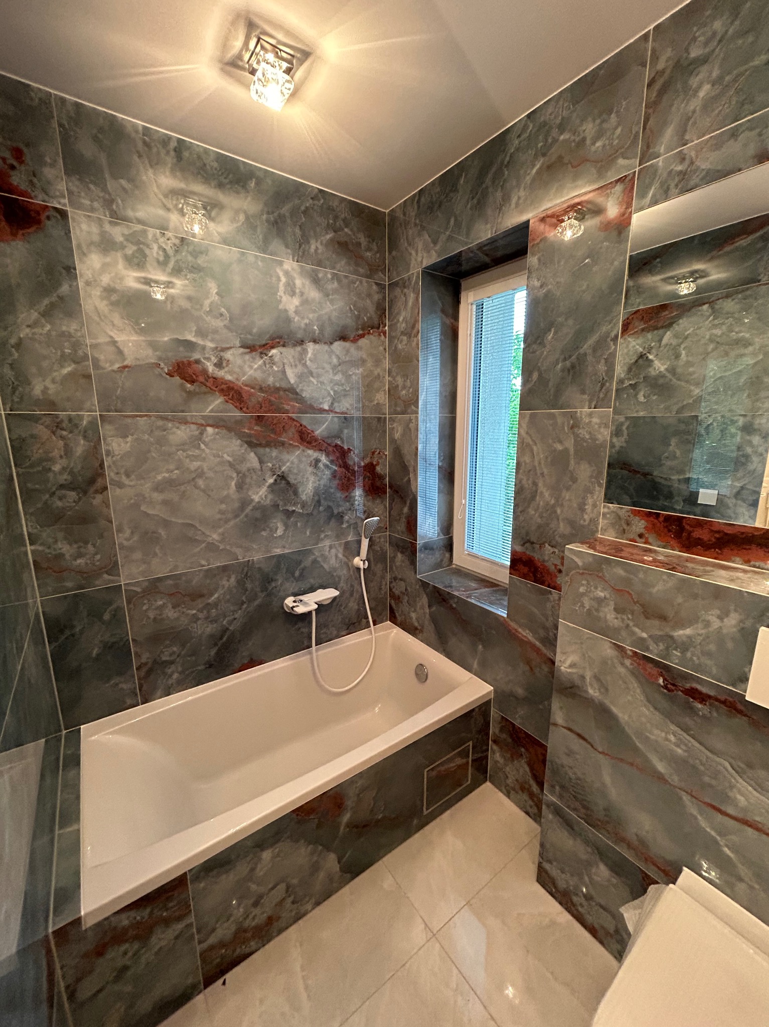 Wykończona łazienka z wanną, szarymi płytkami imitującymi marmur z czerwonymi żyłami, białą armaturą i oknem z żaluzjami.