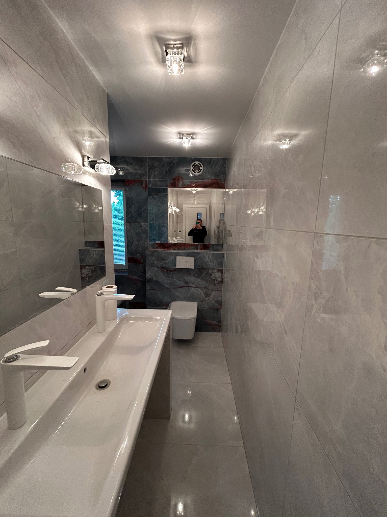 Wąska, wykończona łazienka z szarymi płytkami imitującymi marmur na ścianach i podłodze, biała umywalka nablatowa z nowoczesną baterią, lustro z oświetleniem i odbiciem osoby robiącej zdjęcie...