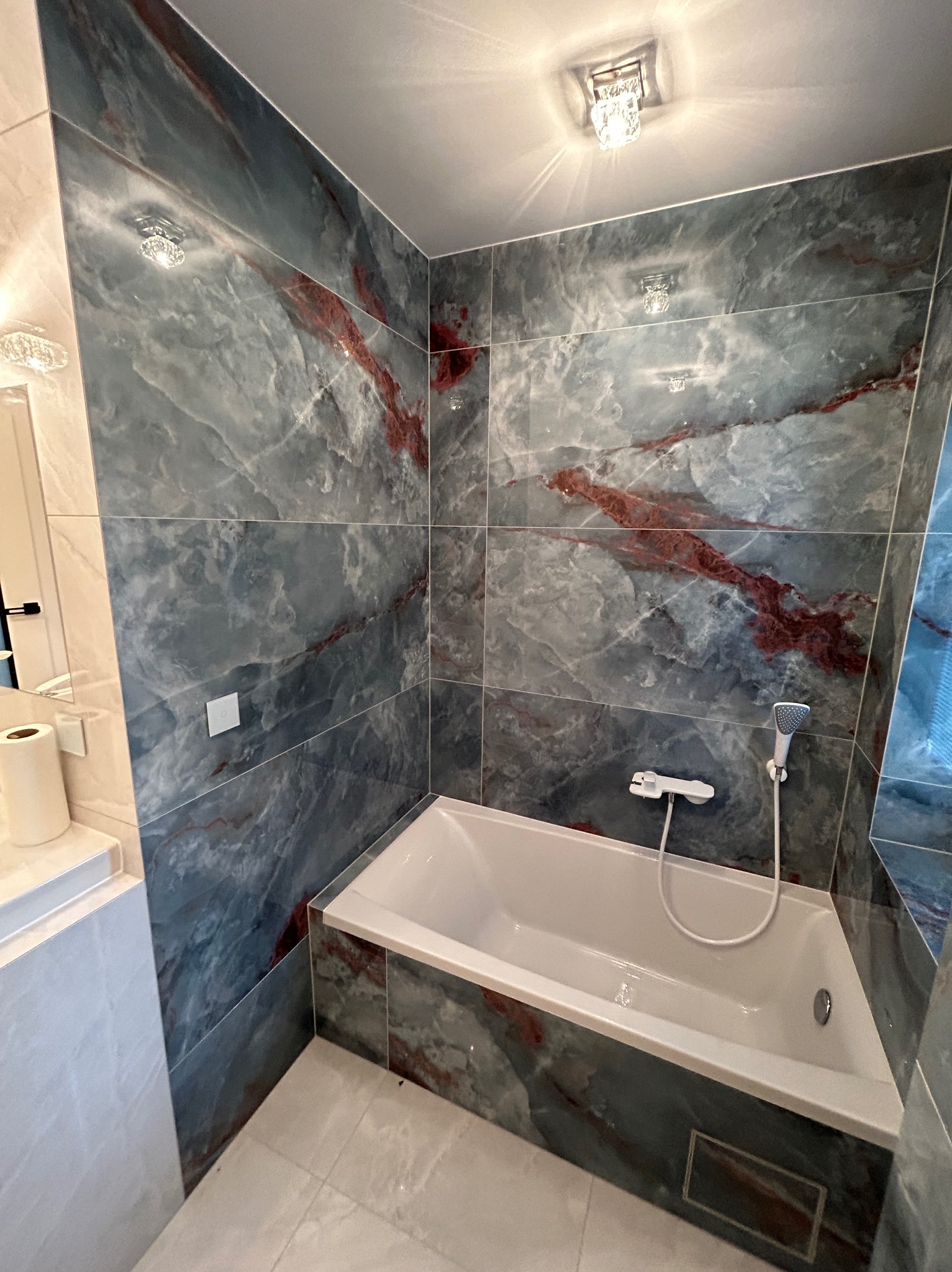 Nowoczesna łazienka z wanną obudowaną płytkami imitującymi marmur w odcieniach szarości z czerwonymi żyłami, podświetlana kryształowymi lampami sufitowymi.