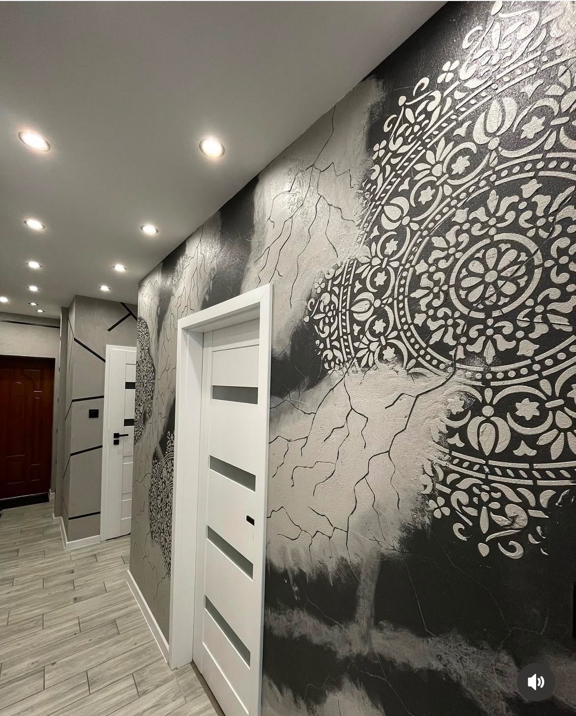 Korytarz z dekoracyjną ścianą w odcieniach szarości z motywem mandali, białe drzwi z poziomymi przeszkleniami i panelowa podłoga w odcieniach szarości.