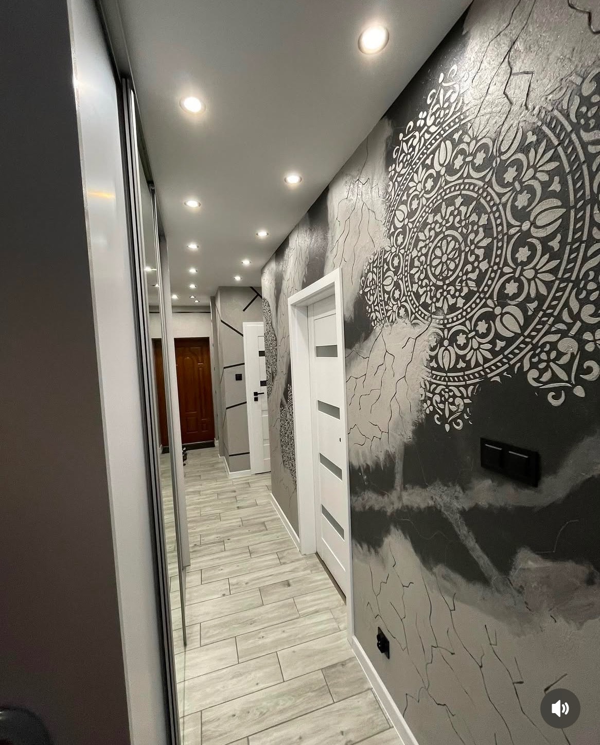 Korytarz z dekoracyjną ścianą w odcieniach szarości, zdobioną białym mandalą i imitacją spękań, jasna podłoga z paneli imitujących drewno, białe drzwi i oświetlenie sufitowe punktowe.