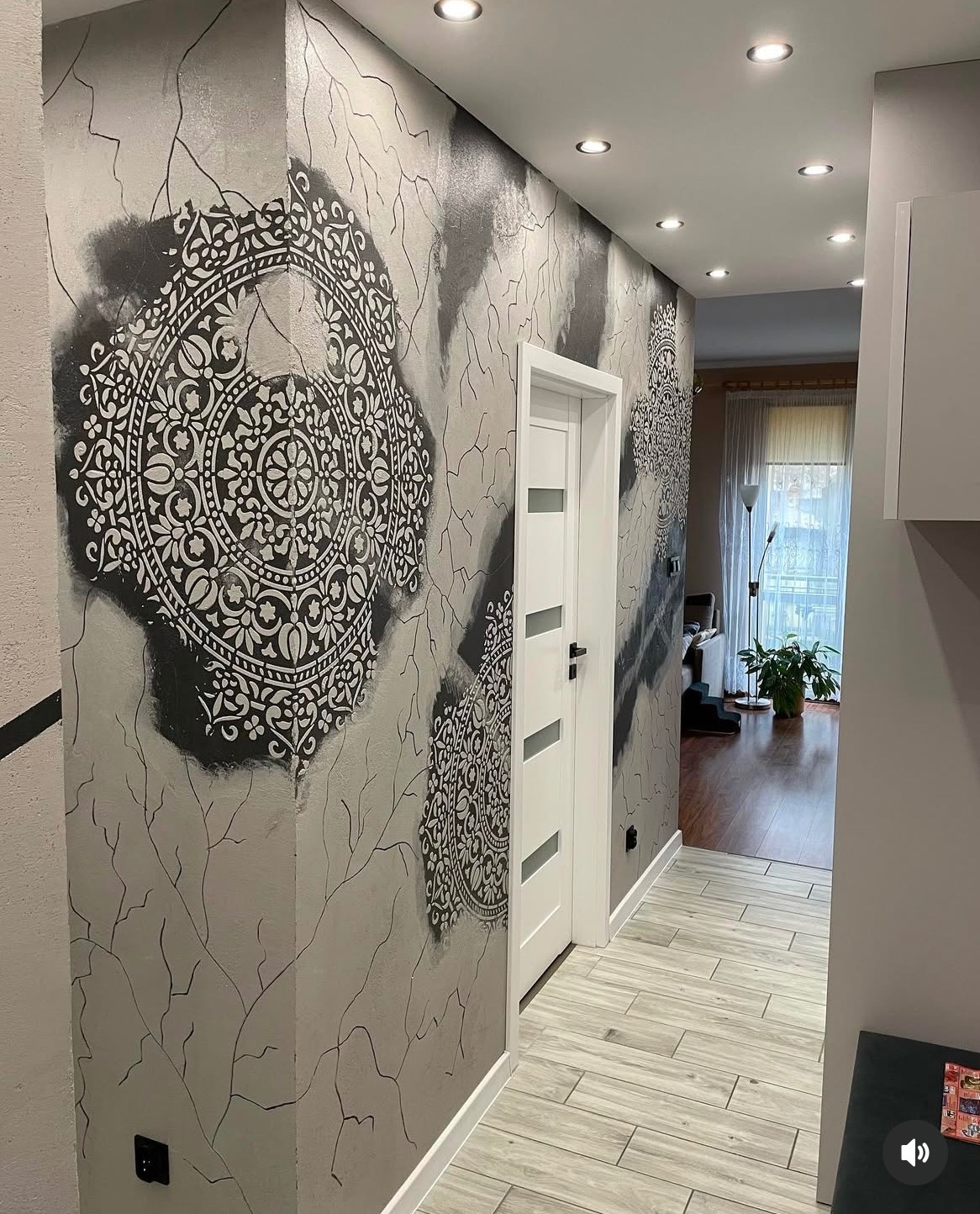 Korytarz z dekoracyjną szarą ścianą z motywem mandali i imitacją spękań, podłoga wyłożona panelami imitującymi drewno, białe drzwi z przeszkleniami, oświetlenie punktowe w suficie.