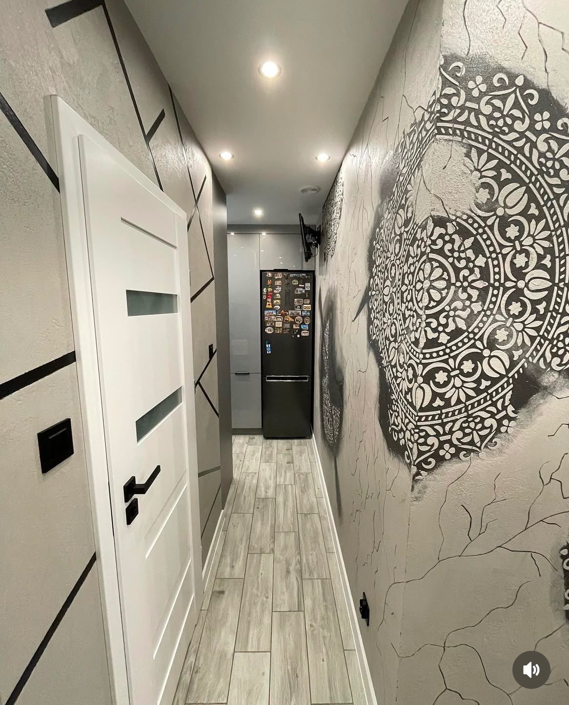 Wąski korytarz z szarą podłogą imitującą drewno, białe drzwi z czarną klamką po lewej stronie, ściana z geometrycznym wzorem i duży ornament w stylu mandali po prawej, w tle widoczna kuchnia...