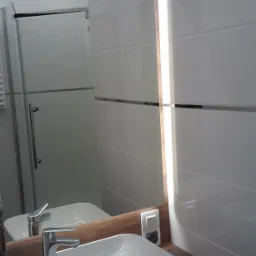 Nowoczesna łazienka z białą, kwadratową umywalką nablatową, chromowaną baterią i podświetlanym lustrem z drewnianą ramą. W tle widoczna kabina prysznicowa z transparentnymi drzwiami.