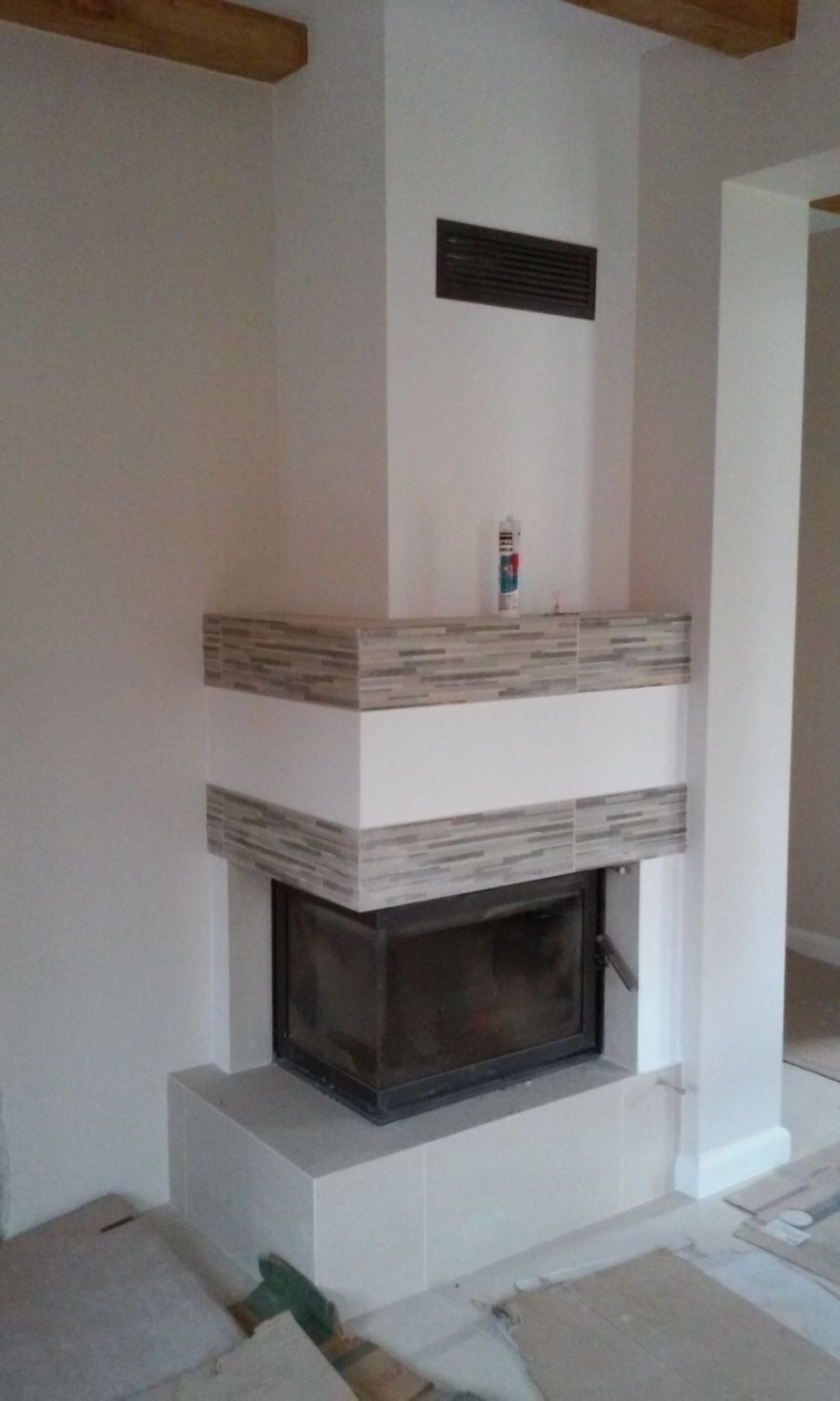 Nowoczesny kominek z czarnym wkładem, obudowany białymi i szarymi płytkami mozaikowymi, z tubą kleju na półce, w trakcie wykańczania.