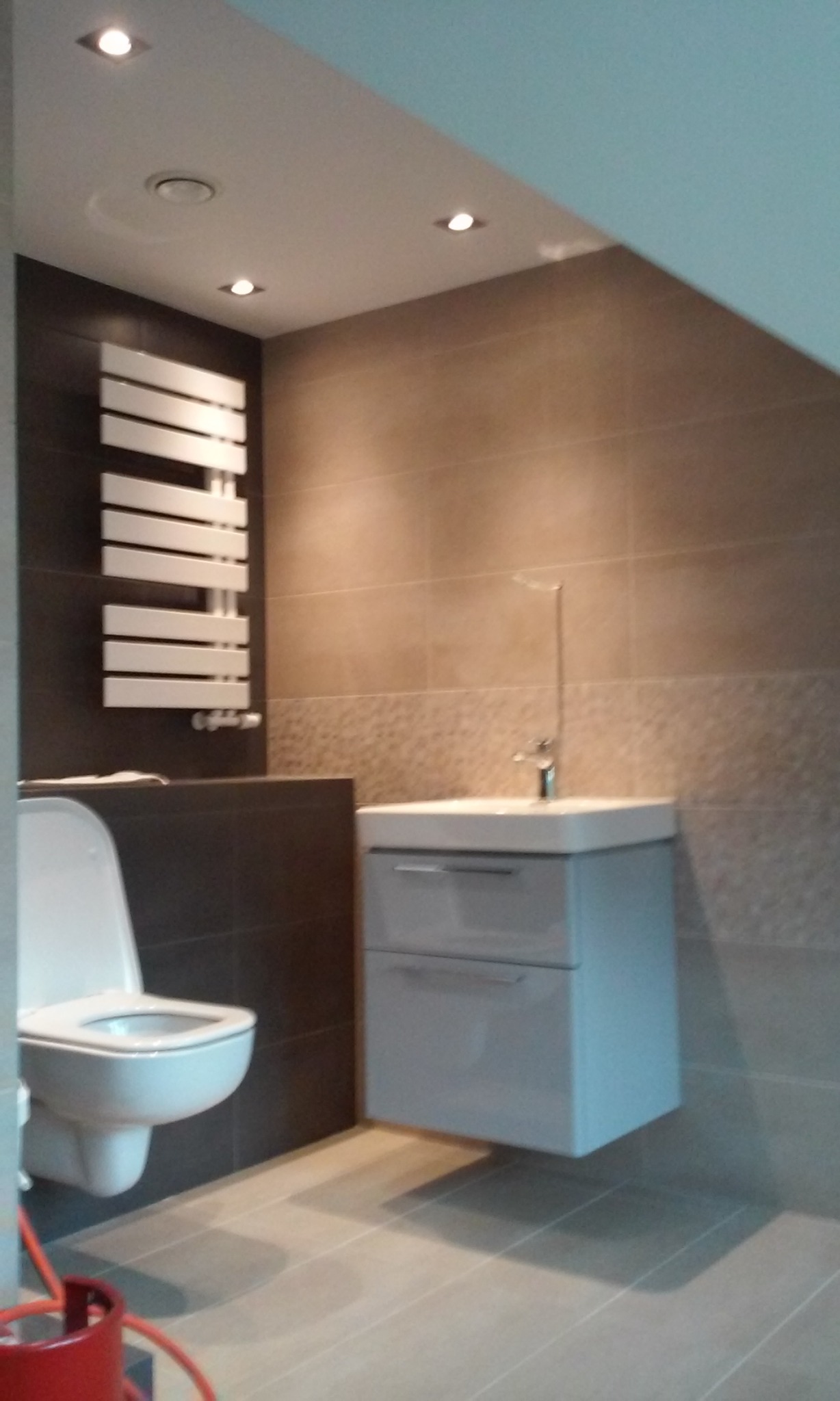 Nowoczesna łazienka z podwieszaną szafką umywalkową, grzejnikiem drabinkowym i toaletą, wykończona brązowo-beżowymi płytkami, oświetlona punktowymi lampami sufitowymi. Widoczny fragment czerwonego...