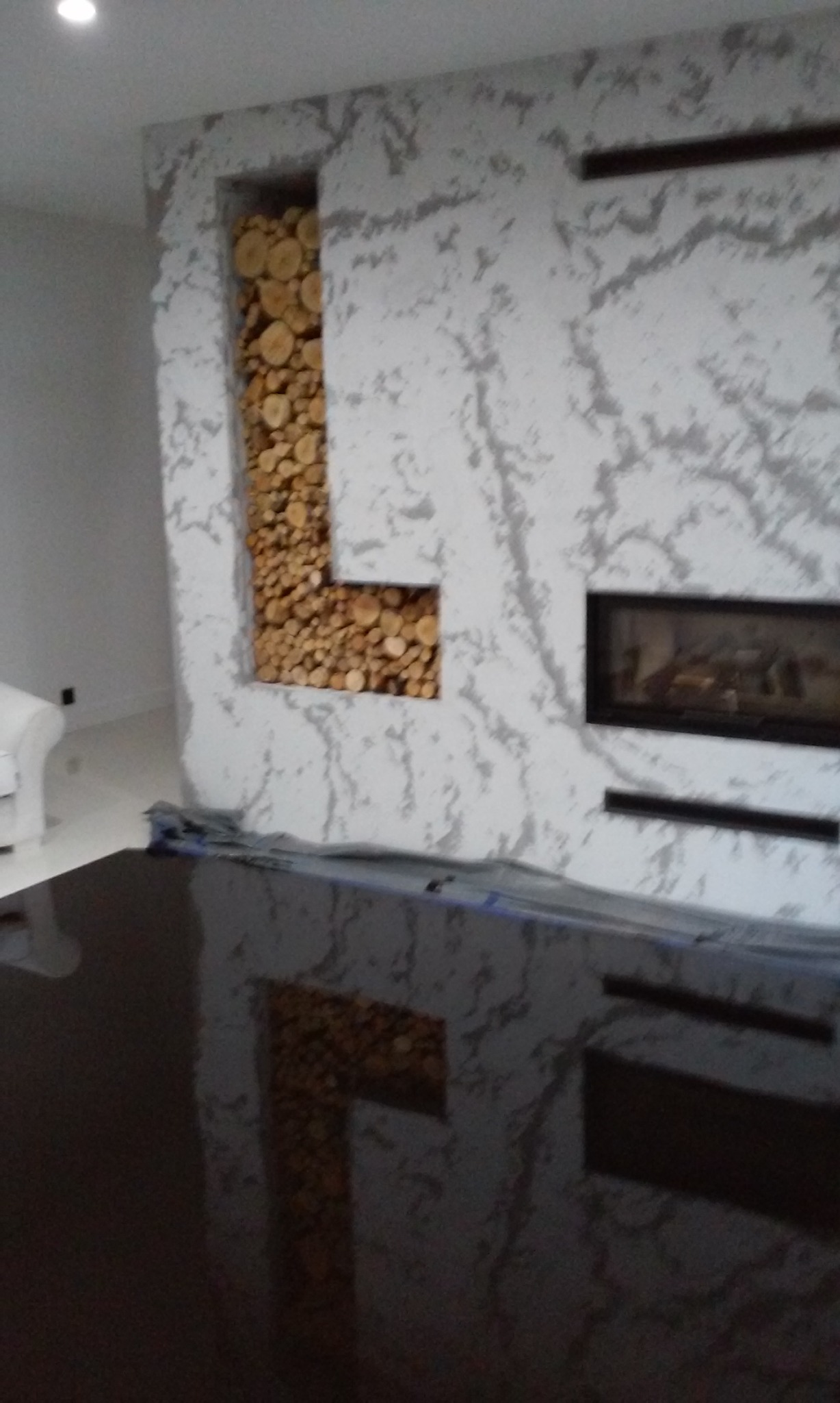 Ściana z dekoracyjnym tynkiem, wnęką wypełnioną drewnem opałowym i wbudowanym kominkiem, częściowo zakryta folią ochronną.