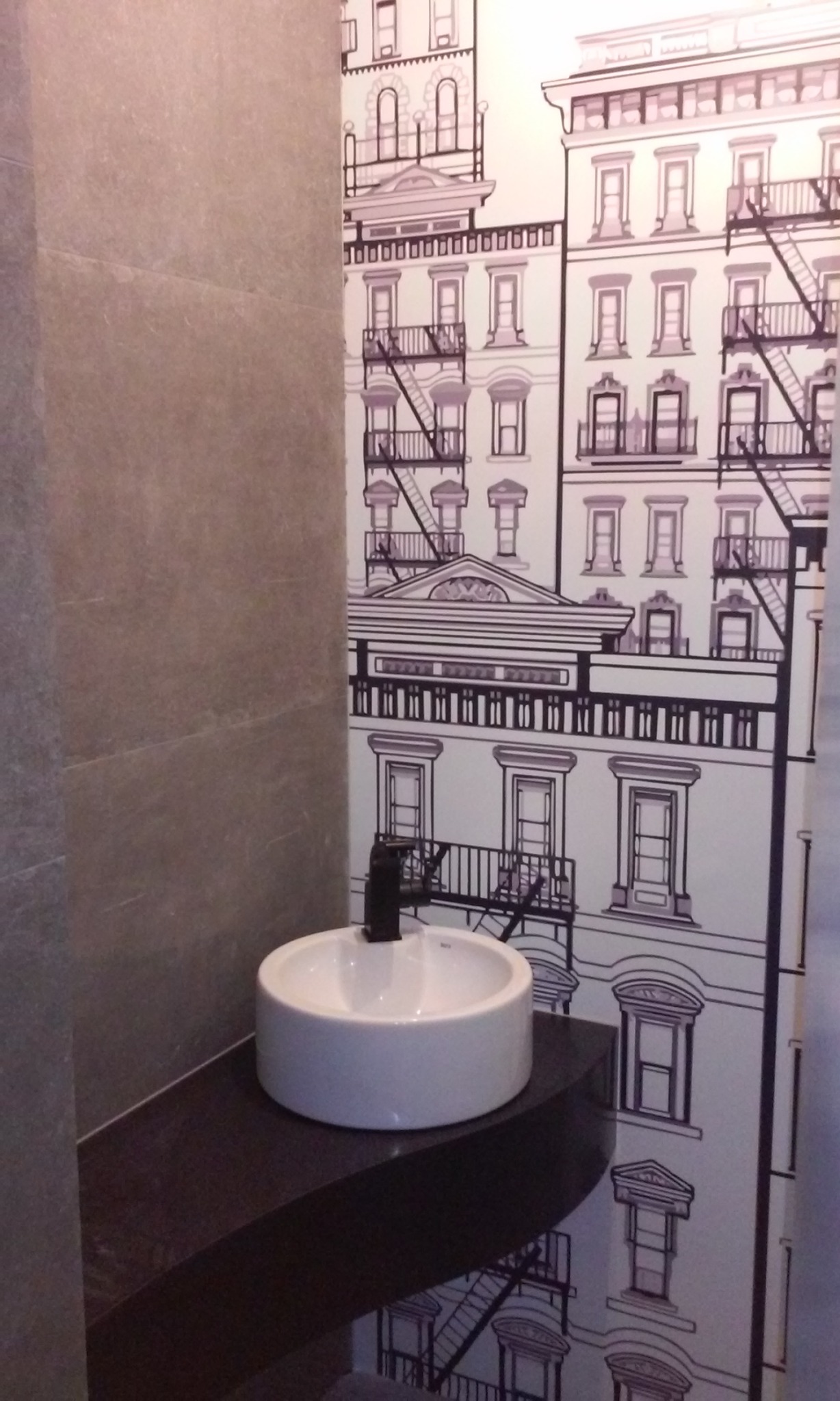 Okrągła, biała umywalka nablatowa z czarną armaturą na ciemnym blacie, na tle ściany z tapetą w rysunkowe kamienice z zewnętrznymi schodami pożarowymi.