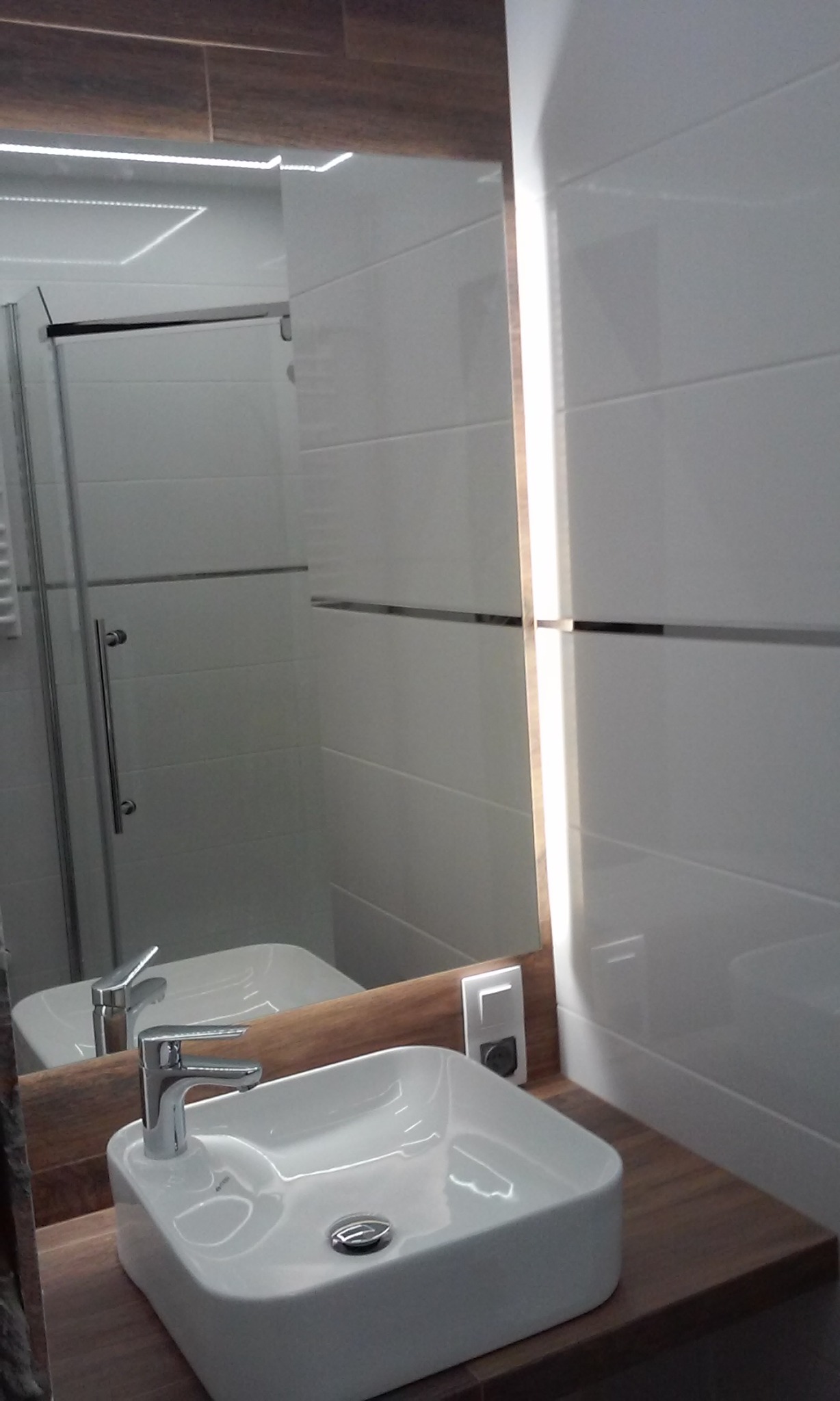 Nowoczesna łazienka z białą, kwadratową umywalką nablatową, chromowaną baterią i podświetlanym lustrem z drewnianą ramą. W tle widoczna kabina prysznicowa z transparentnymi drzwiami.