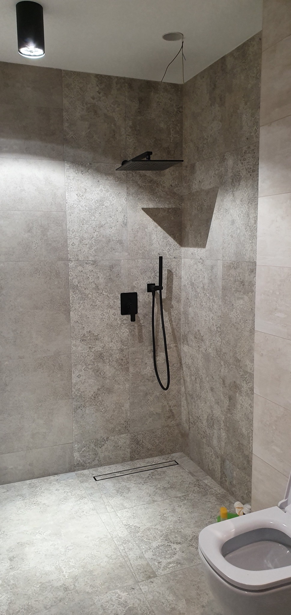 Nowoczesna kabina prysznicowa z deszczownicą i czarną armaturą, wykończona płytkami w odcieniach szarości, widoczny odpływ liniowy i fragment toalety, surowy minimalizm.