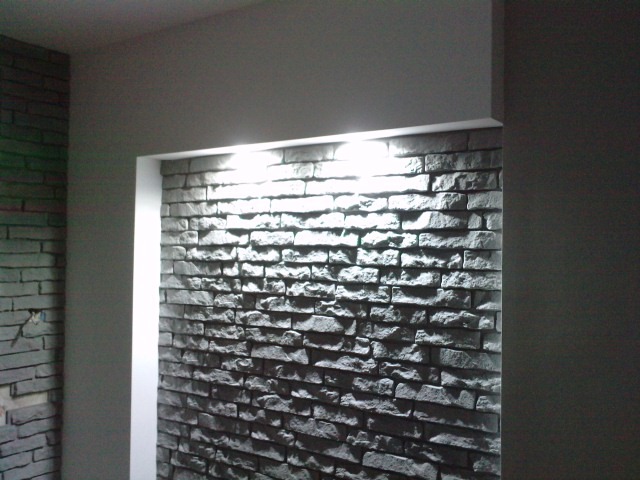 Fragment ściany z dekoracyjnej, szarej cegły z podświetleniem LED w górnej części wnęki, widoczny fragment drugiej ściany z cegły w trakcie montażu.
