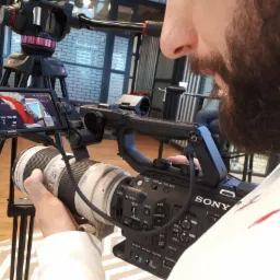 Operator kamery Sony z brodą, ustawia ostrość obiektywu podczas filmowania, widoczny podgląd nagrywanego materiału na ekranie kamery, statyw Manfrotto w tle.