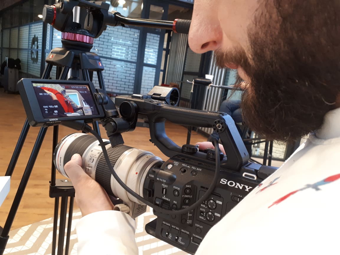 Operator kamery Sony z brodą, ustawia ostrość obiektywu podczas filmowania, widoczny podgląd nagrywanego materiału na ekranie kamery, statyw Manfrotto w tle.