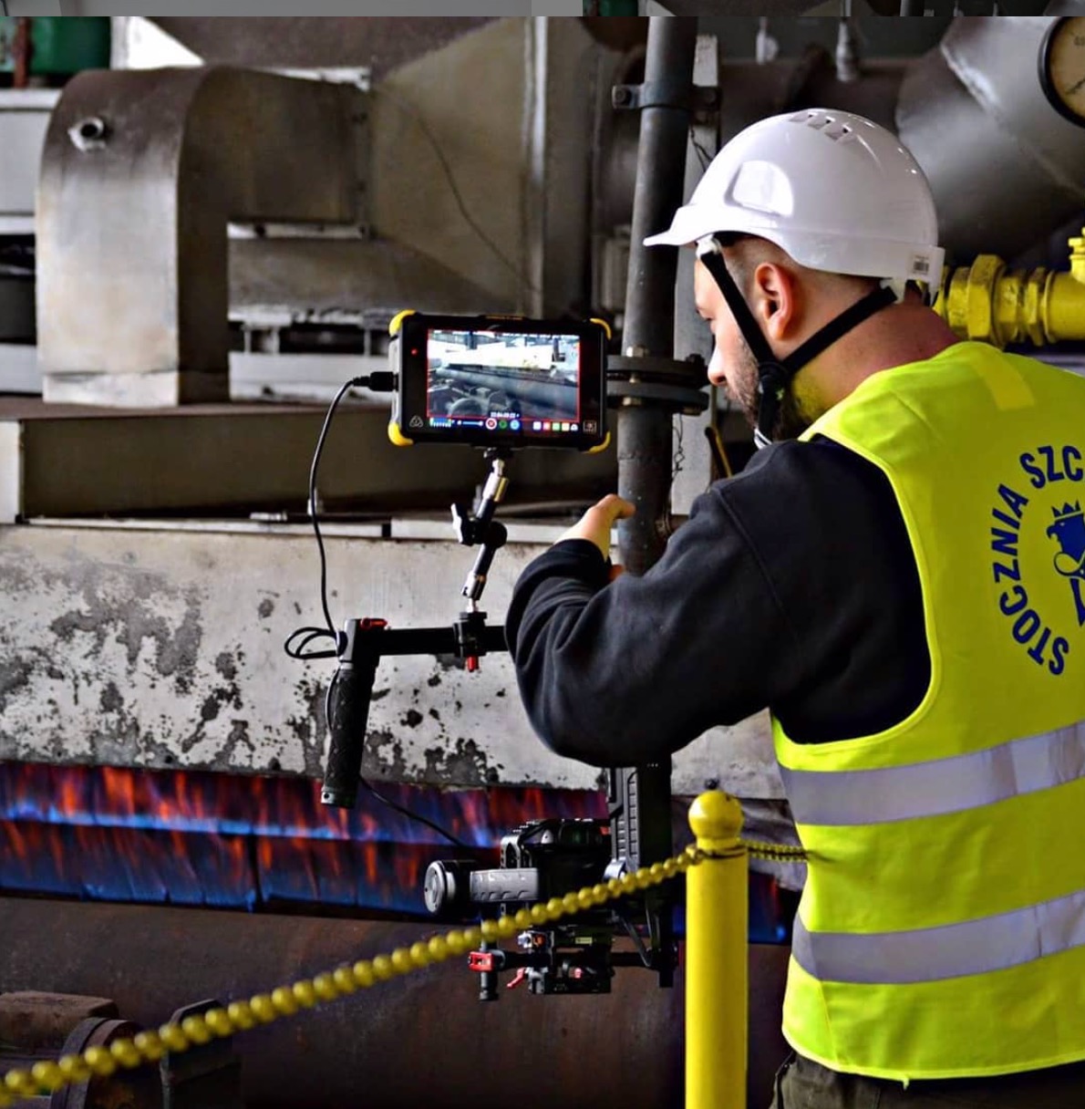 Operator kamery w kasku i kamizelce odblaskowej filmuje wnętrze hali Stoczni Szczecińskiej, w tle widoczne płomienie palnika.