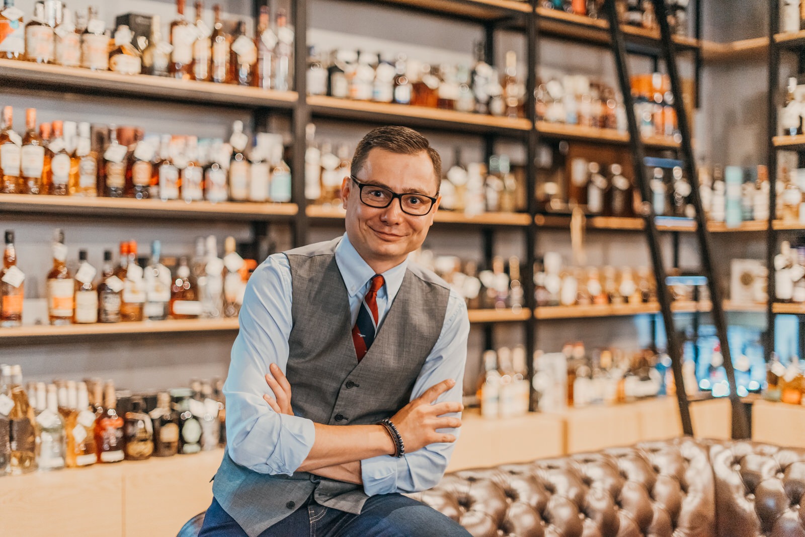 Uśmiechnięty mężczyzna w okularach i kamizelce siedzi na skórzanej sofie na tle półek z butelkami alkoholu.