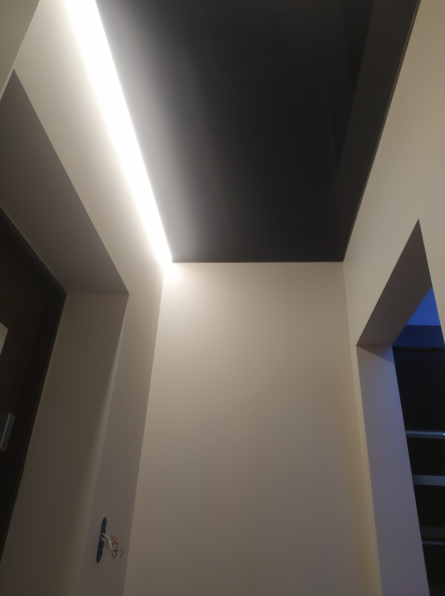 Czarne lustrzane odbicie sufitu napinanego z podświetleniem LED wzdłuż krawędzi, widoczne niedokończone gniazdko elektryczne na ścianie.