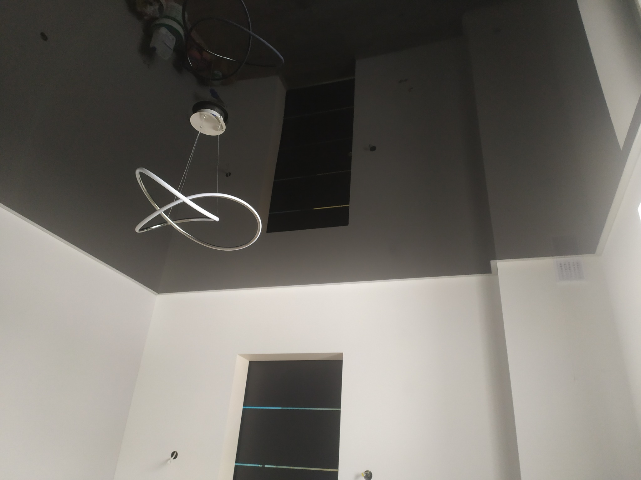 Ciemnoszary, błyszczący sufit z zamontowaną nowoczesną lampą w kształcie dwóch przecinających się okręgów, widoczne białe ściany i fragment czarnej wnęki w suficie.