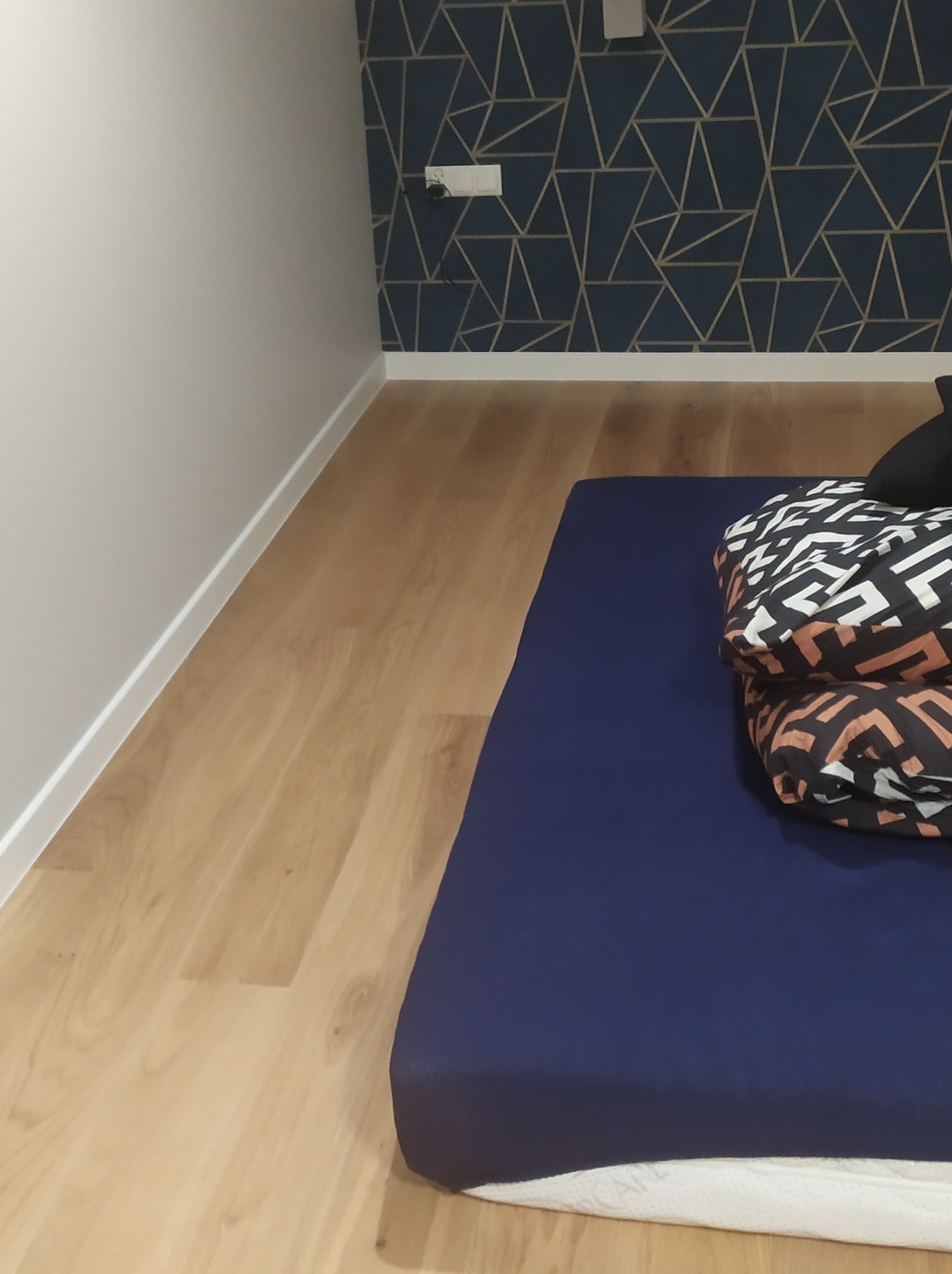 Fragment pokoju we Frysztaku po remoncie: podłoga z paneli, ściana pomalowana na biało z listwą przypodłogową, ściana z tapetą w geometryczne wzory, na podłodze materac i poduszki.
