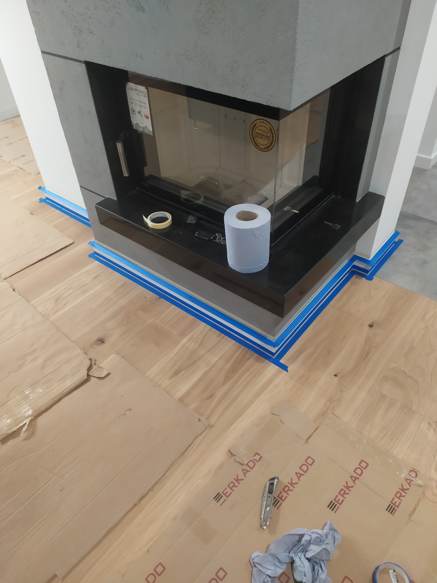 Montaż kominka: zabezpieczona podłoga kartonem z logo, taśma malarska na styku podłogi i obudowy kominka, rolka papieru toaletowego na czarnym blacie kominka.