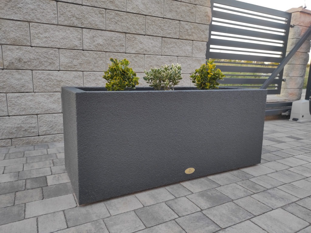 Długa, prostokątna, szara donica z trzema małymi krzewami o zielono-żółtych liściach, ustawiona na szarej kostce brukowej, z fragmentem muru z jasnoszarej cegły i czarną, nowoczesną bramą w tle.