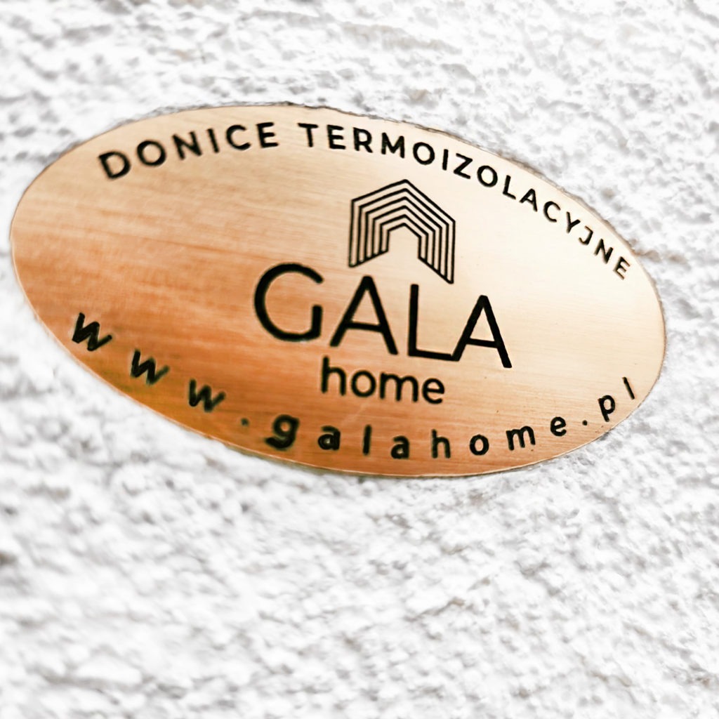 Miedziana tabliczka z logo i adresem strony internetowej 'GALA home' na tle białej, chropowatej powierzchni. Napis 'DONICE TERMOIZOLACYJNE' umieszczony wzdłuż górnej krawędzi.