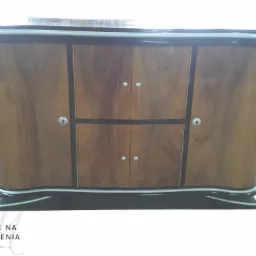 Komoda w stylu art déco z forniru orzechowego, z czterema drzwiczkami i chromowanymi uchwytami, wykończona czarnym lakierem na obrzeżach i podstawie, z logo producenta w lewym dolnym rogu.