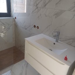 Usługi Ogólnobudowlane Daniel Kudzbalski - Nowoczesna łazienka z marmurowymi płytkami, białą szafką pod umywalką i szklaną kabiną prysznicową. Widoczne przygotowane punkty instalacyjne.