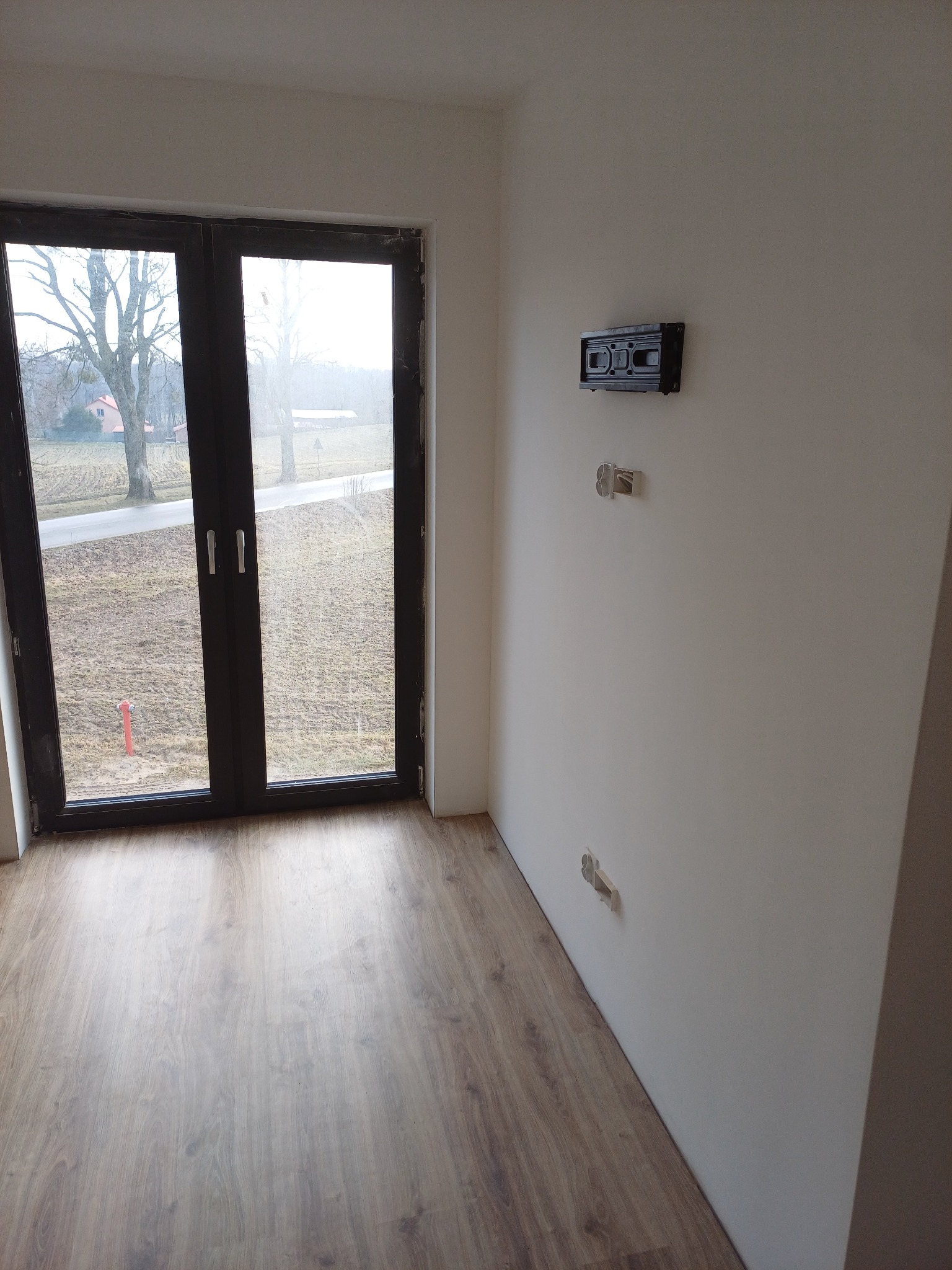 Pomalowane na biało wnętrze pokoju z panelami podłogowymi, widoczne gniazdka elektryczne i uchwyt na telewizor na ścianie. Za oknem widok na zewnątrz.