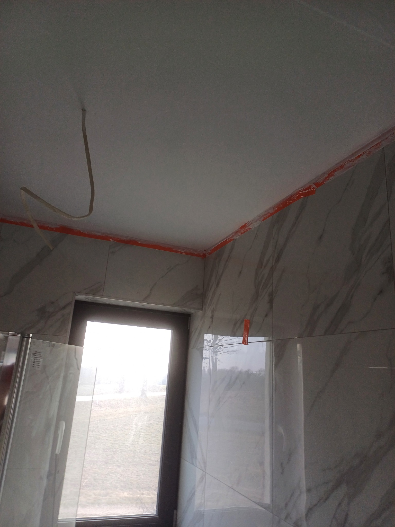 Łazienka w trakcie wykańczania: marmurowe płytki na ścianach, zabezpieczone folią, okno z widokiem na zewnątrz, wystający kabel z sufitu. Widoczne fragmenty kabiny prysznicowej.