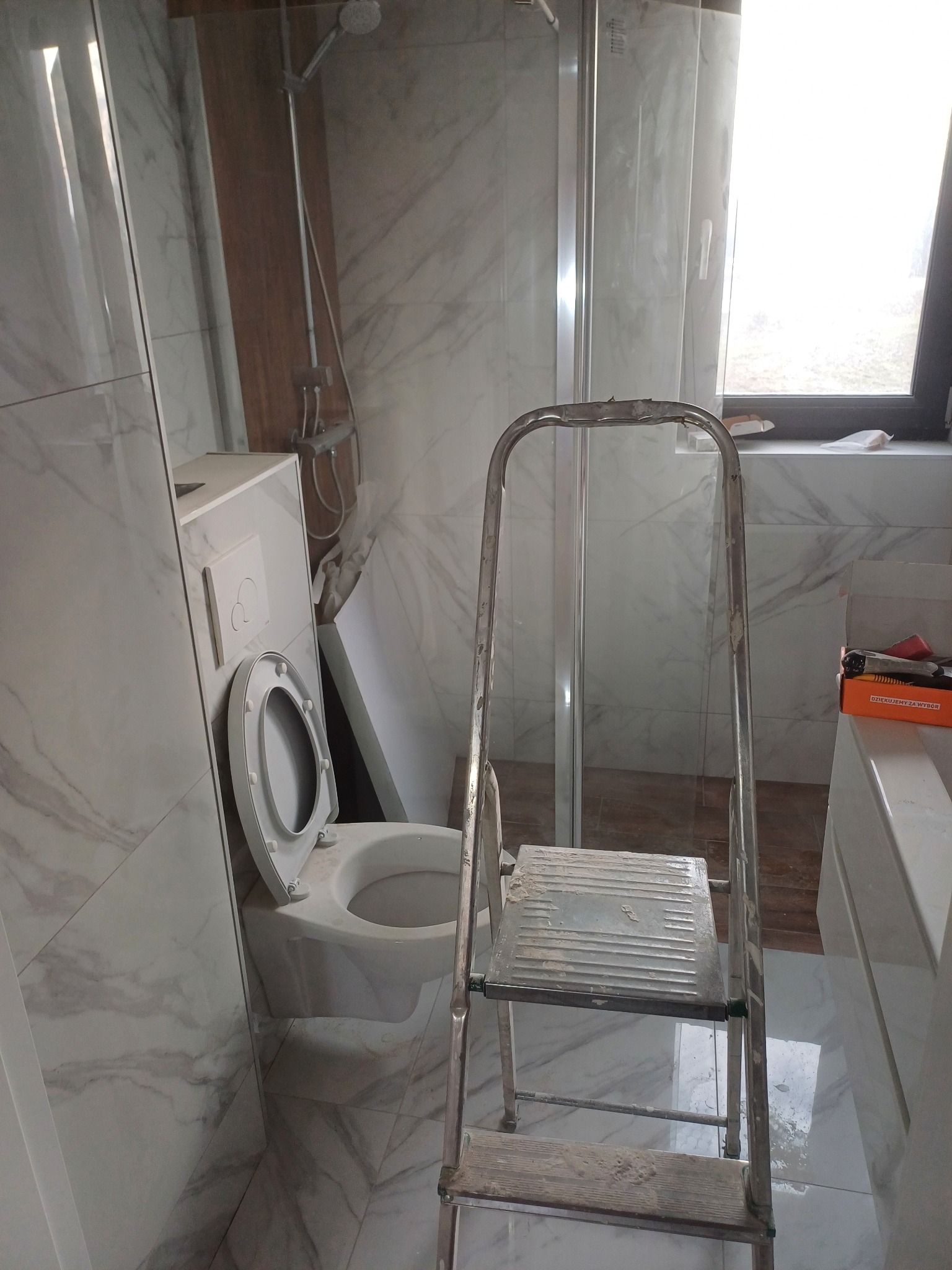 Remont łazienki w trakcie: widoczna toaleta podwieszana, kabina prysznicowa i drabina. Marmurowe płytki na ścianach. Etap prac wykończeniowych.