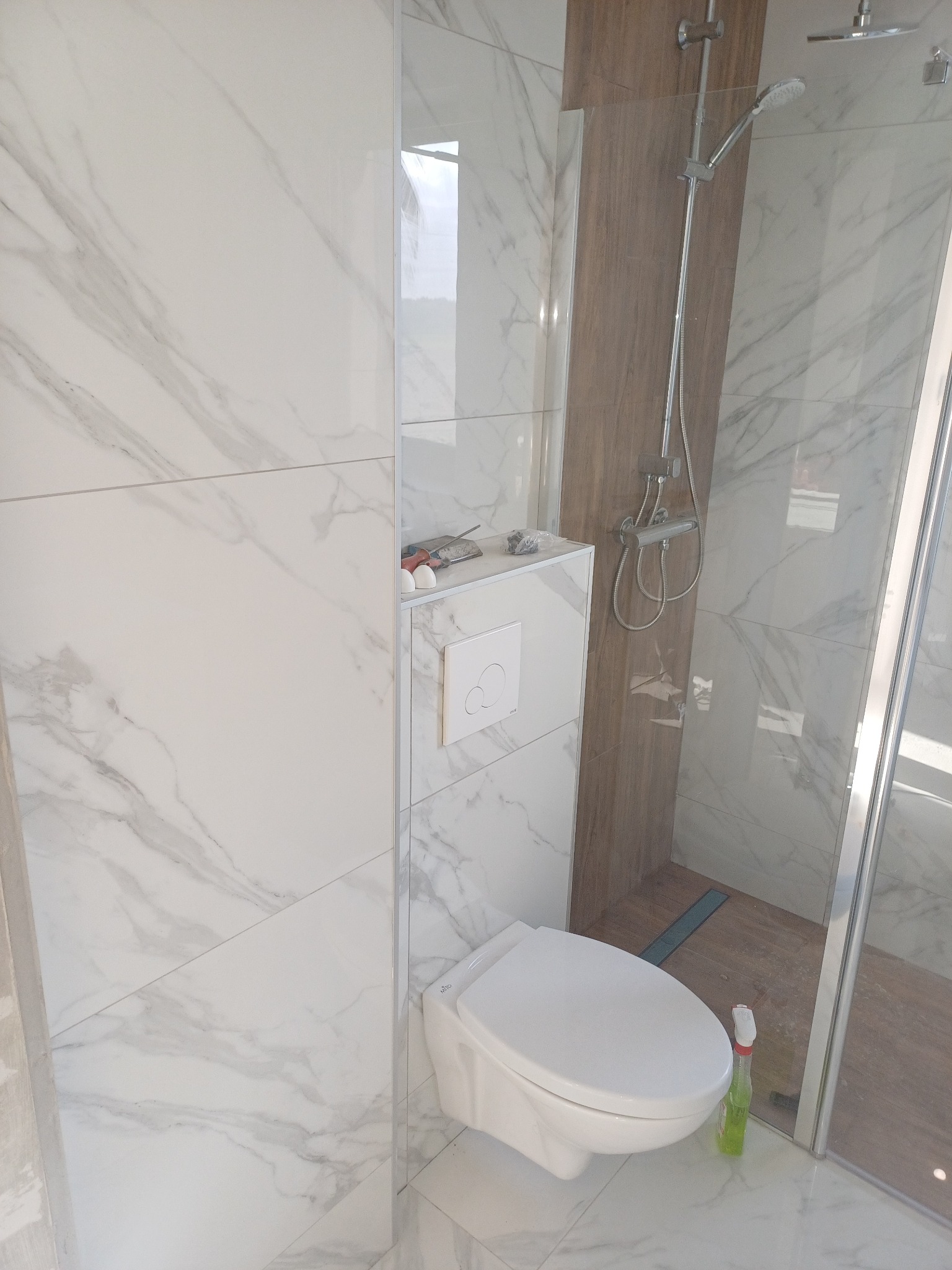 Wykończona łazienka z kabiną prysznicową i toaletą w zabudowie. Białe płytki imitujące marmur na ścianach i podłodze. Widoczny fragment kabiny i armatura.