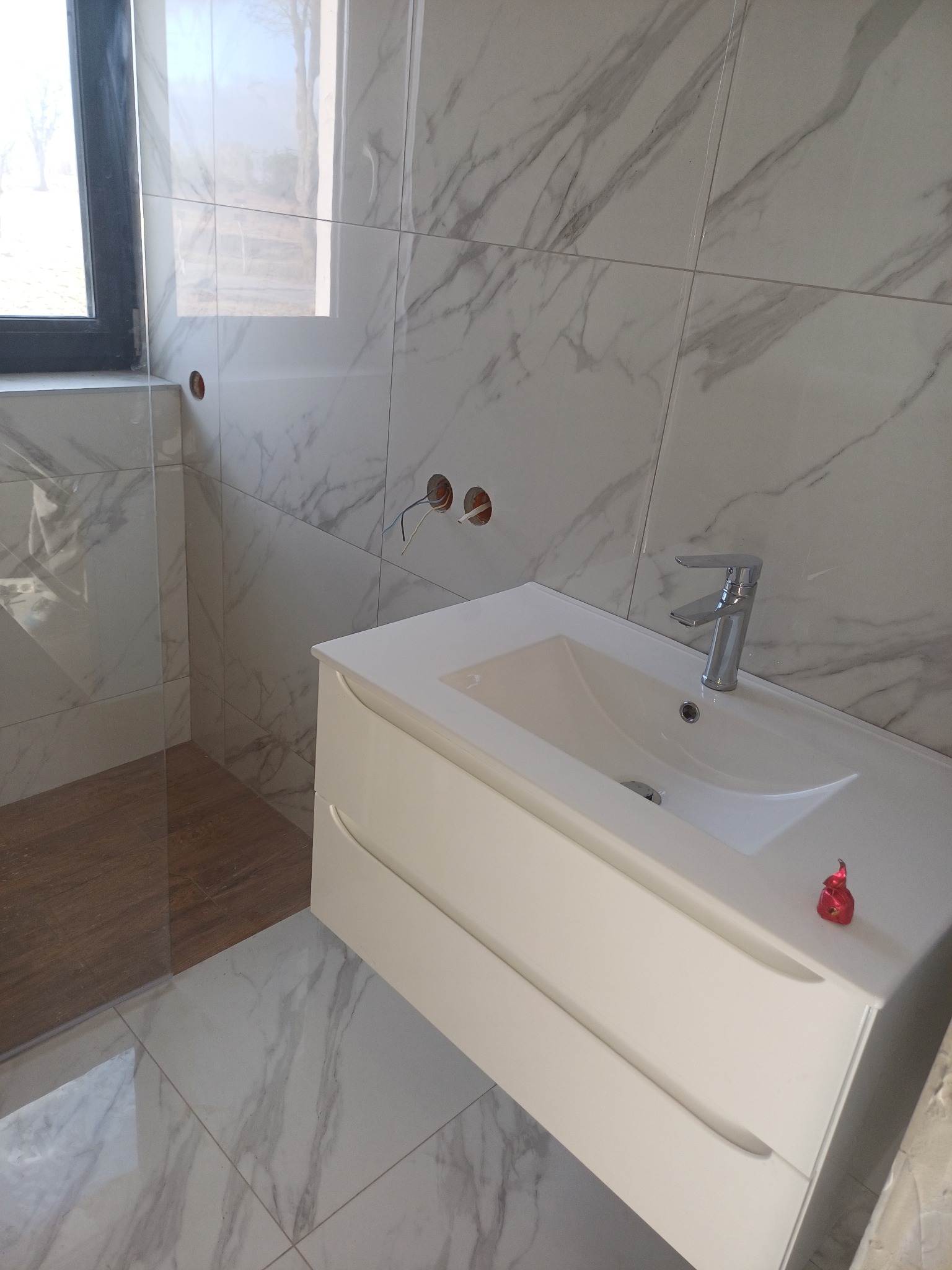 Nowoczesna łazienka z marmurowymi płytkami, białą szafką pod umywalką i szklaną kabiną prysznicową. Widoczne przygotowane punkty instalacyjne.