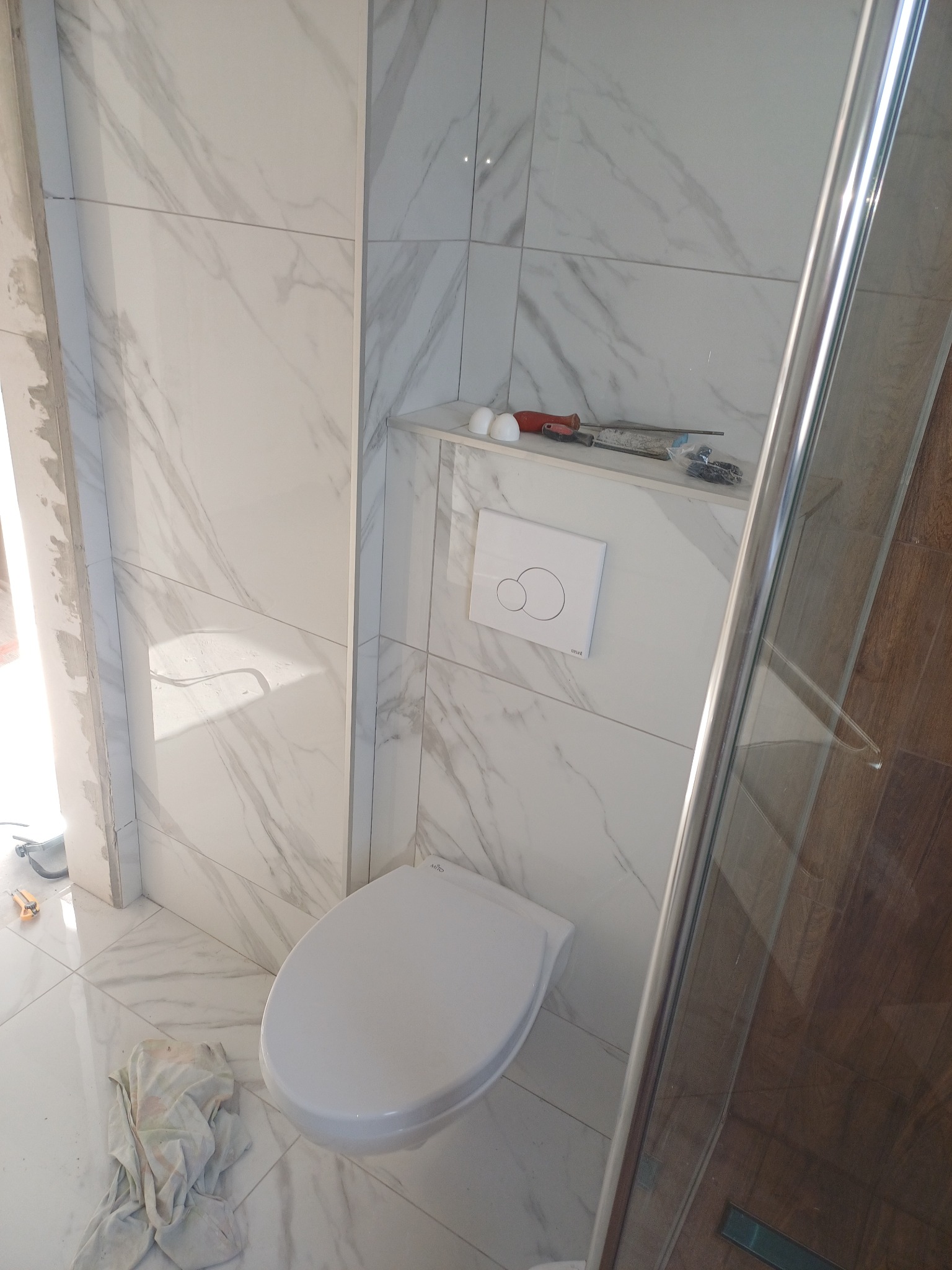 Biała toaleta wisząca w wykończonej łazience z marmurowymi płytkami. Widoczne narzędzia na półce i fragment kabiny prysznicowej. Ukończony etap prac.