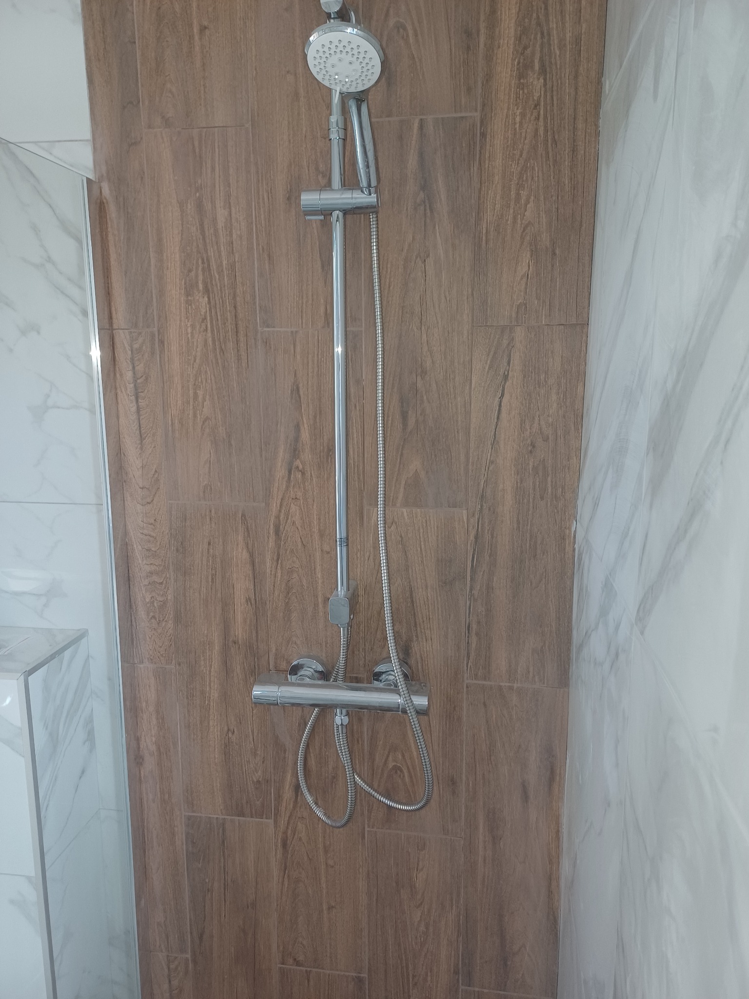 Nowoczesny zestaw prysznicowy z deszczownicą na ścianie wyłożonej brązowymi płytkami imitującymi drewno, obok kabina z marmurowymi płytkami. Minimalistyczny design.