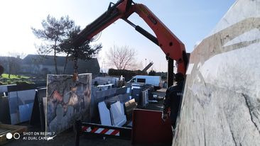 Czerwony dźwig HDS podnosi płytę z jasnego kamienia na placu składowym, widoczne inne płyty kamienne i budynek w tle.