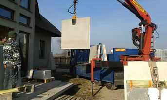 Rozładunek betonowych płyt za pomocą dźwigu samochodowego w Blachowni. Na pierwszym planie płyta zawieszona na haku dźwigu, w tle budynek mieszkalny i paleta z materiałami budowlanymi.