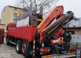 Czerwona ciężarówka z żurawiem hydraulicznym Palfinger, zaparkowana na tle budynku mieszkalnego i ogrodzenia. Widoczny numer rejestracyjny pojazdu.