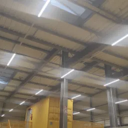 Wnętrze hali przemysłowej z zamontowanym oświetleniem LED, widok z dołu na sufit z widocznymi belkami konstrukcyjnymi, świetlikami i żółtym kontenerem w tle.