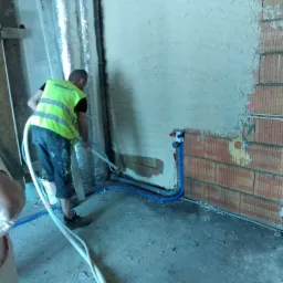 Pracownik w kamizelce odblaskowej nanosi tynk maszynowy na ścianę z cegły dziurawki, widoczne rury instalacyjne w ścianie i przewody tynkarskie na podłodze.