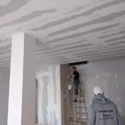 Nakładanie pierwszej warstwy gładzi na suficie w nowym mieszkaniu; widoczne pasy świeżo nałożonego materiału, pracownik na drabinie z kielnią i wiaderkiem, w tle klimatyzator, oraz osoba w bluzie...