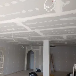 Wnętrze w trakcie prac wykończeniowych: zamontowane płyty gipsowo-kartonowe na suficie i ścianach, częściowo zaszpachlowane łączenia, widoczne przewody elektryczne, drabiny i narzędzia budowlane.