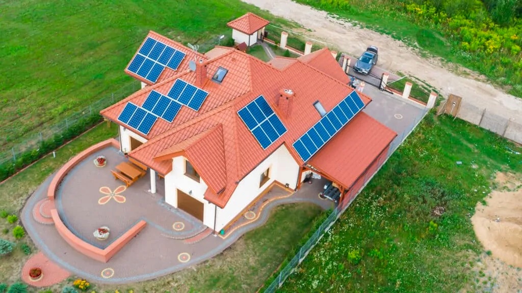 Widok z góry na dom jednorodzinny z czerwoną dachówką i panelami słonecznymi na dachu, z zadbanym ogrodem, patio z kwiatowym wzorem i zaparkowanym samochodem w tle.