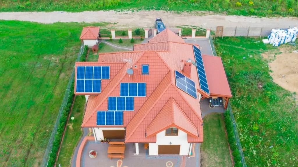 Widok z góry na dom z czerwoną dachówką i panelami słonecznymi na dachu, otoczony zielonym trawnikiem i ogrodzeniem. W tle zaparkowany samochód.