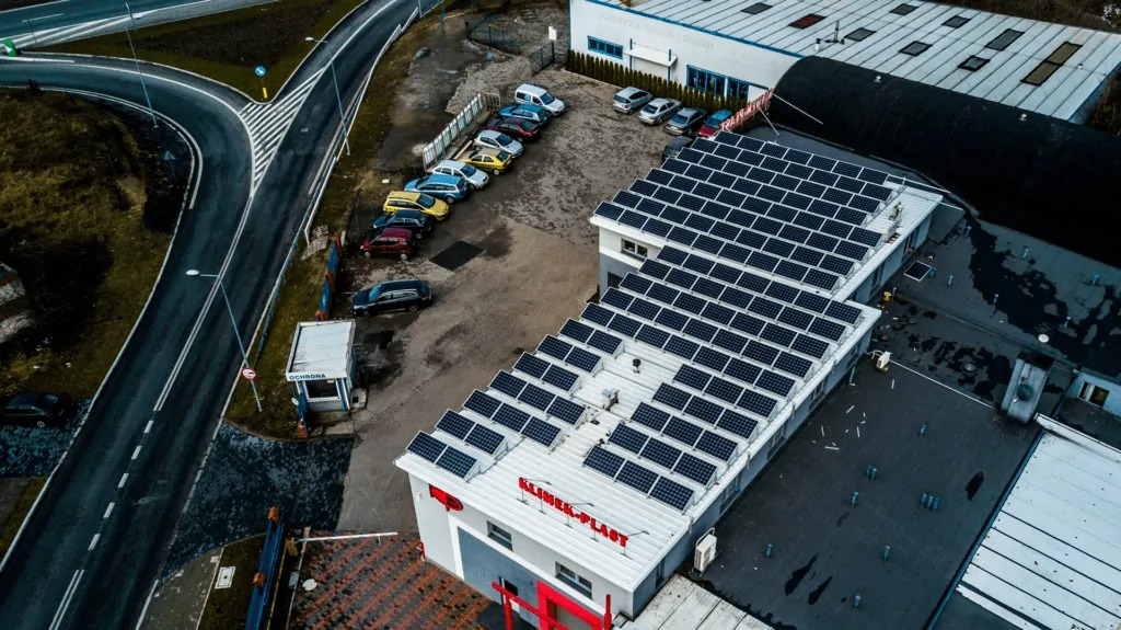 Widok z góry na budynek firmy Klimex-Plast z zainstalowanymi panelami słonecznymi na dachu, obok parking z samochodami i droga dojazdowa.