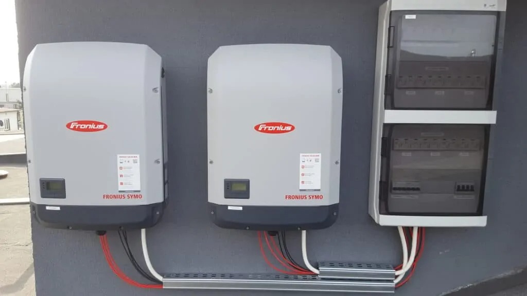 Dwa falowniki Fronius Symo zamontowane obok skrzynki elektrycznej na szarej ścianie, widoczne kable w listwach przypodłogowych.