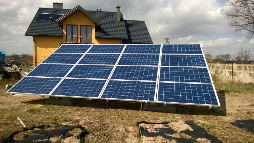 Zestaw paneli słonecznych na betonowych podstawach przed żółtym domem z ciemnym dachem, widoczne również dodatkowe panele na dachu domu.