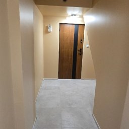 Bogdan Bróg - Beżowy korytarz po remoncie z szarą podłogą z płytek i drewnianymi drzwiami z czarnym paskiem. Widok z perspektywy.