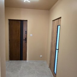 Bogdan Bróg - Beżowy przedpokój z dwiema drewnianymi drzwiami, szarą podłogą z płytek i nowoczesnym oświetleniem sufitowym.