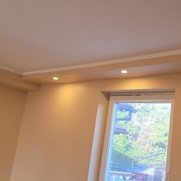 Bogdan Bróg - Jasne wnętrze po remoncie: beżowe ściany, biały sufit z oświetleniem punktowym i okno z widokiem na zieleń, wykończone listwy przysufitowe.